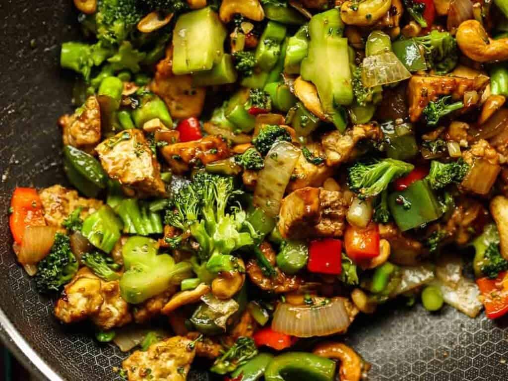 Close-up of a stir-fry featuring a mix of vibrant vegetables including broccoli, bell peppers, and onions, along with tofu and cashews, all seasoned and cooked in a skillet.